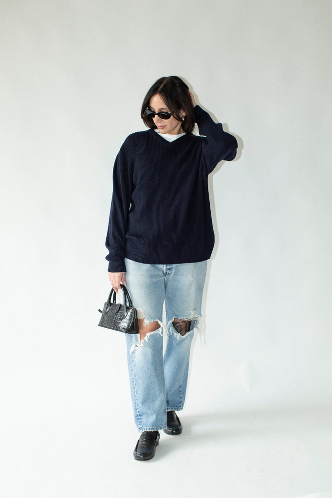Person wearing a navy sweater and light blue jeans with a small handbag on a white background