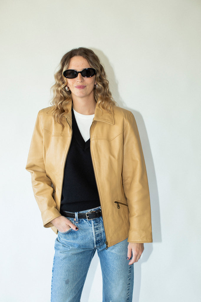 Woman wearing a tan leather jacket, black top, and blue jeans against a white background