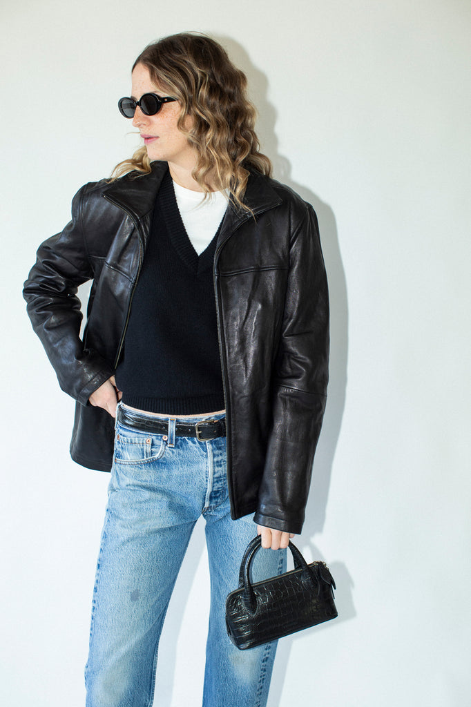 Person wearing a black leather jacket, black sweater, blue jeans, and holding a black handbag against a white background.