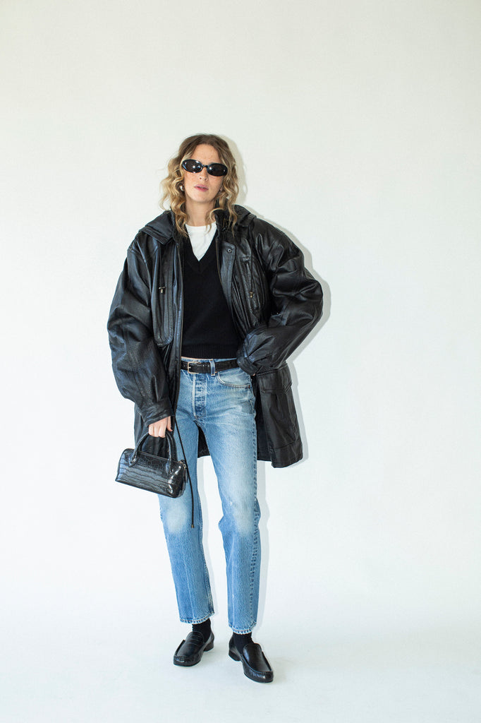 Person wearing a black coat, black shirt, blue jeans, and black shoes on a white background
