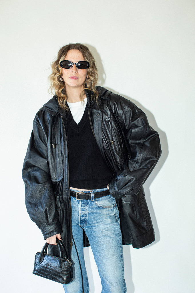 Person wearing a black leather jacket, black shirt, blue jeans, and sunglasses on a white background