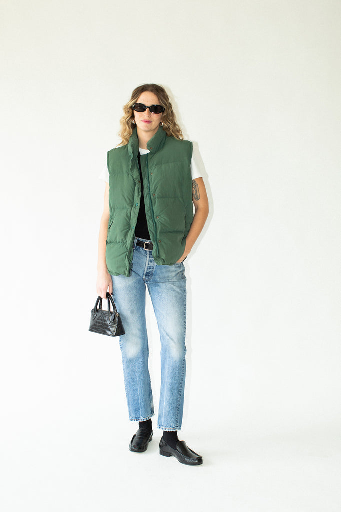 Person wearing a green vest, white shirt, blue jeans, and black shoes on a white background