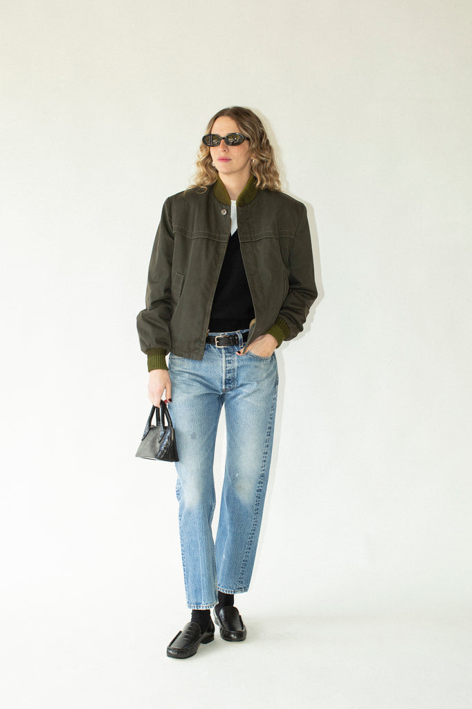 Person wearing a green jacket, black shirt, light blue jeans, and black shoes on a white background