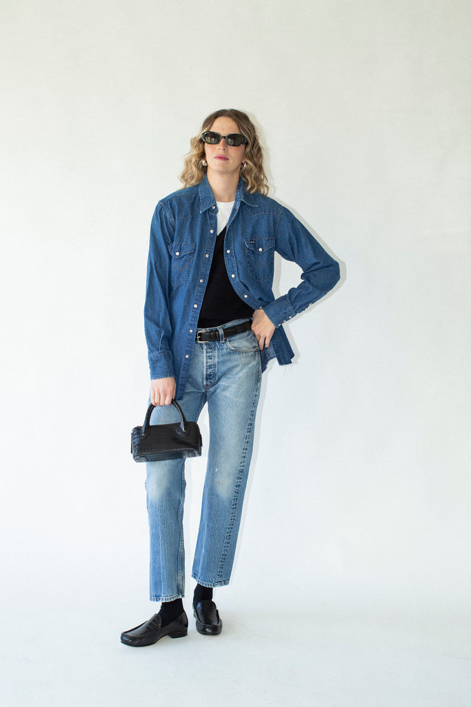 Person wearing a blue denim shirt, jeans, and black shoes, holding a black handbag against a white background.