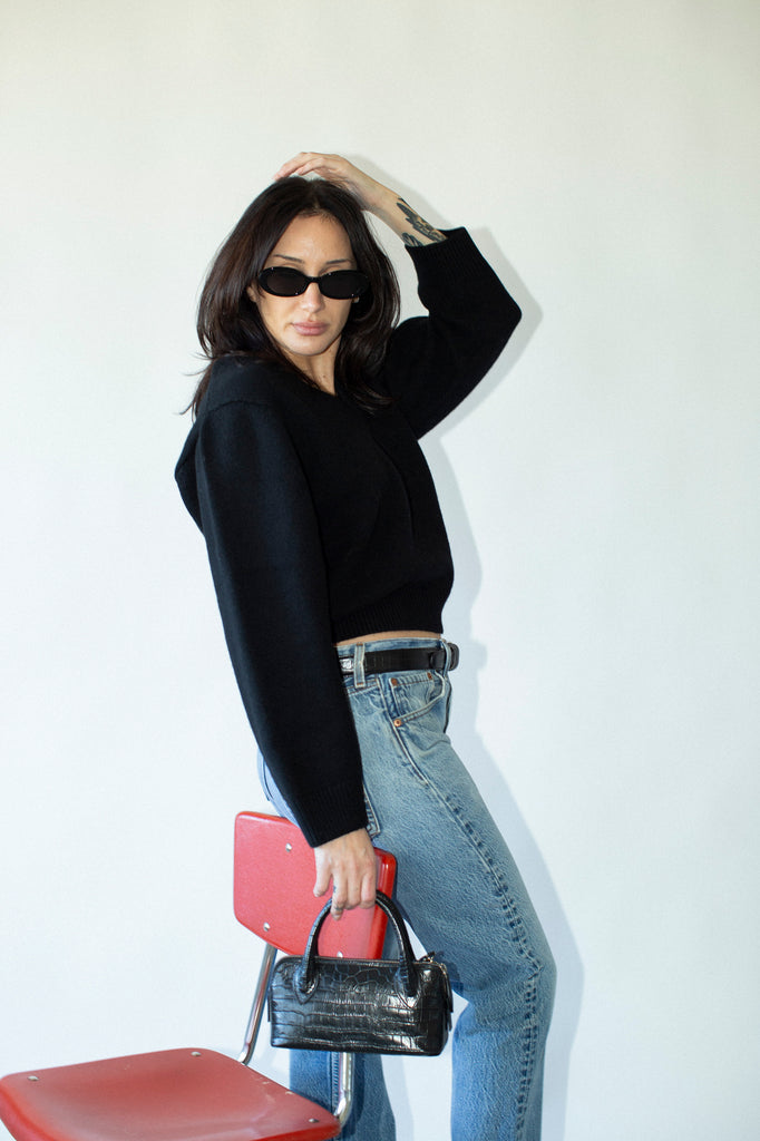 Woman in black top and blue jeans holding a handbag against a white background