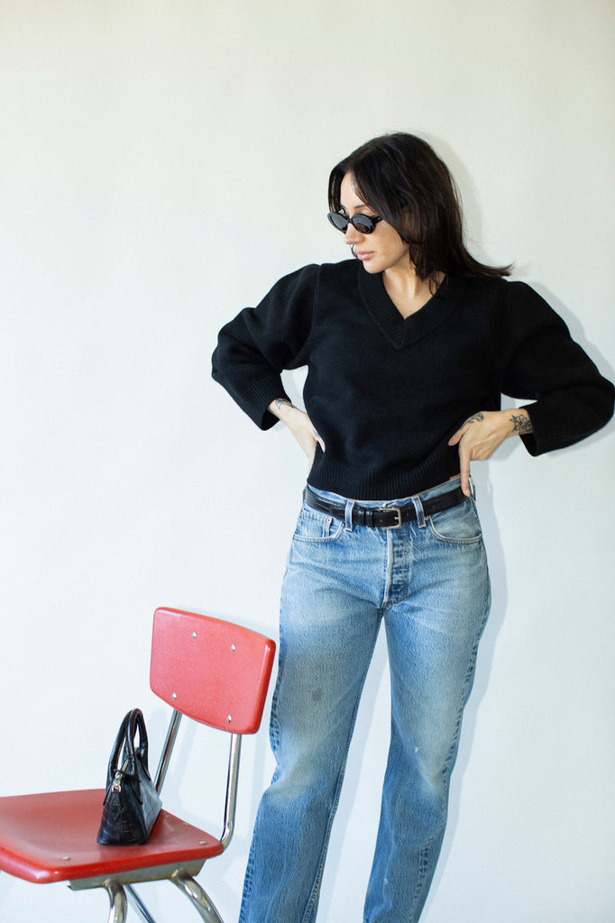 Woman in black sweater and blue jeans standing next to a red chair against a white wall.