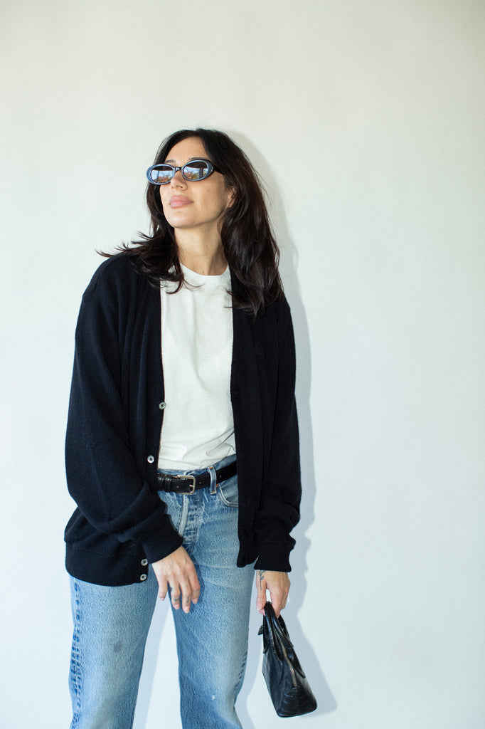 Woman wearing sunglasses, black cardigan, white shirt, blue jeans, and holding a black handbag against a white wall.