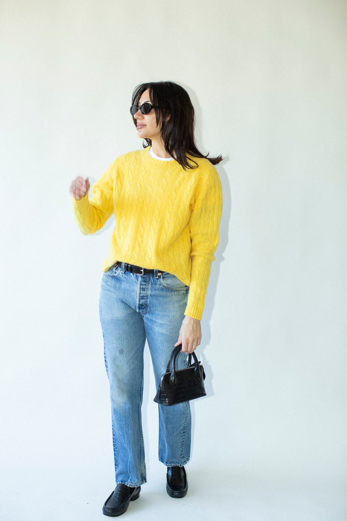 Woman wearing a yellow sweater, blue jeans, and black shoes against a white background