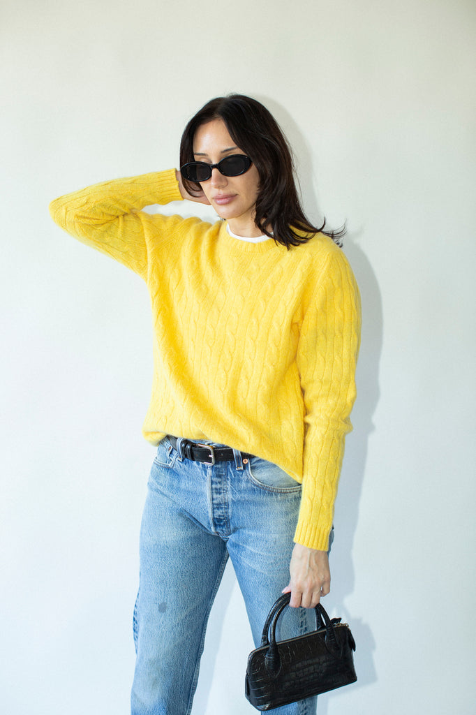 Woman wearing a yellow sweater and blue jeans, holding a black handbag against a white background
