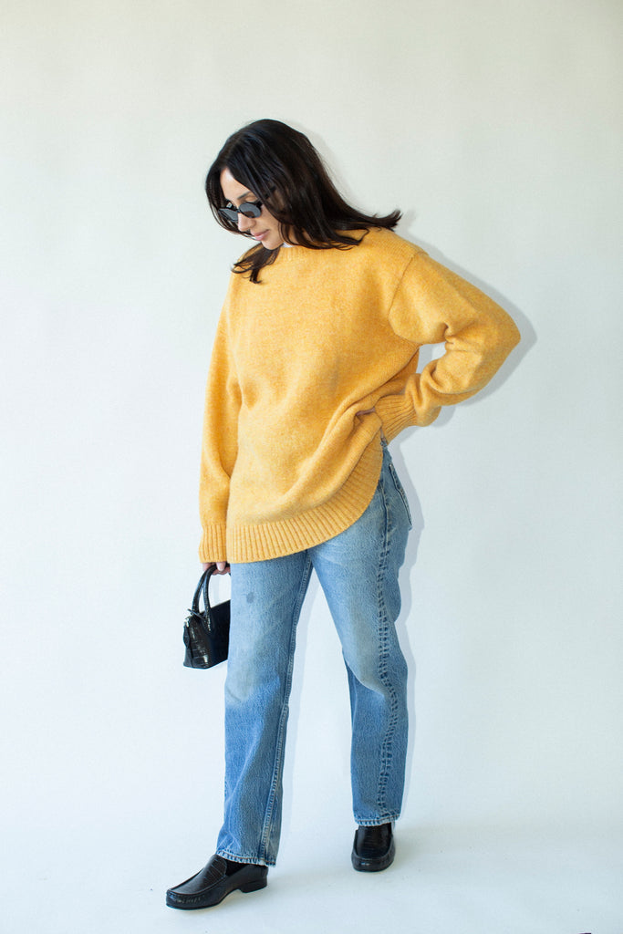 Woman wearing a mustard yellow sweater and blue jeans against a white background