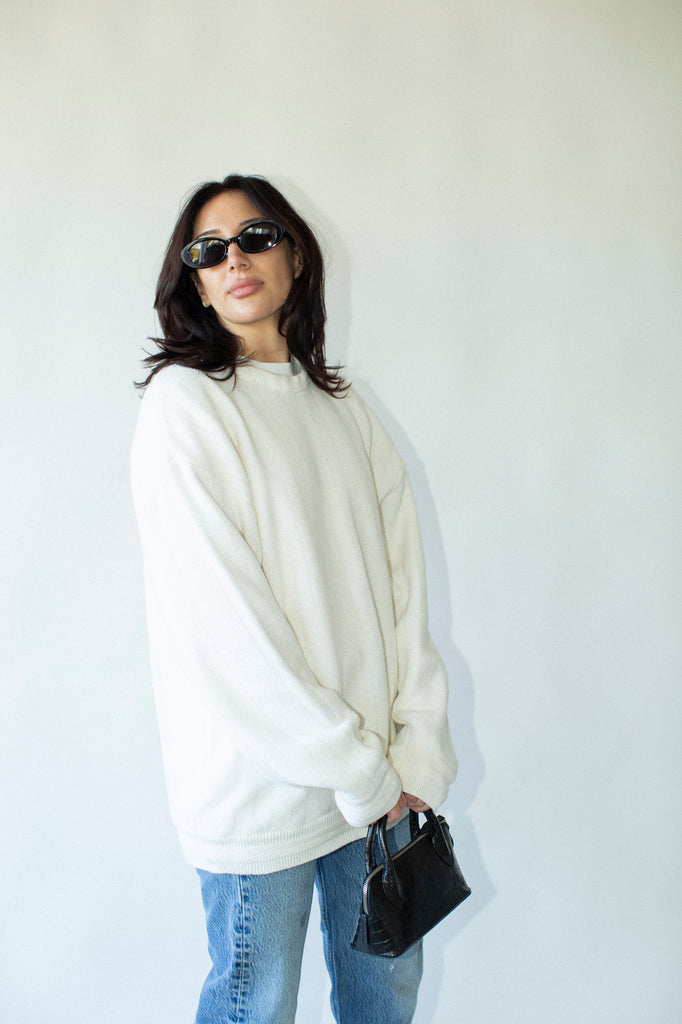 Woman wearing a white oversized sweater and blue jeans, holding a black handbag against a plain background.