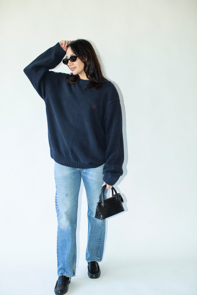 Woman wearing a navy sweater and light blue jeans, holding a black handbag against a white background