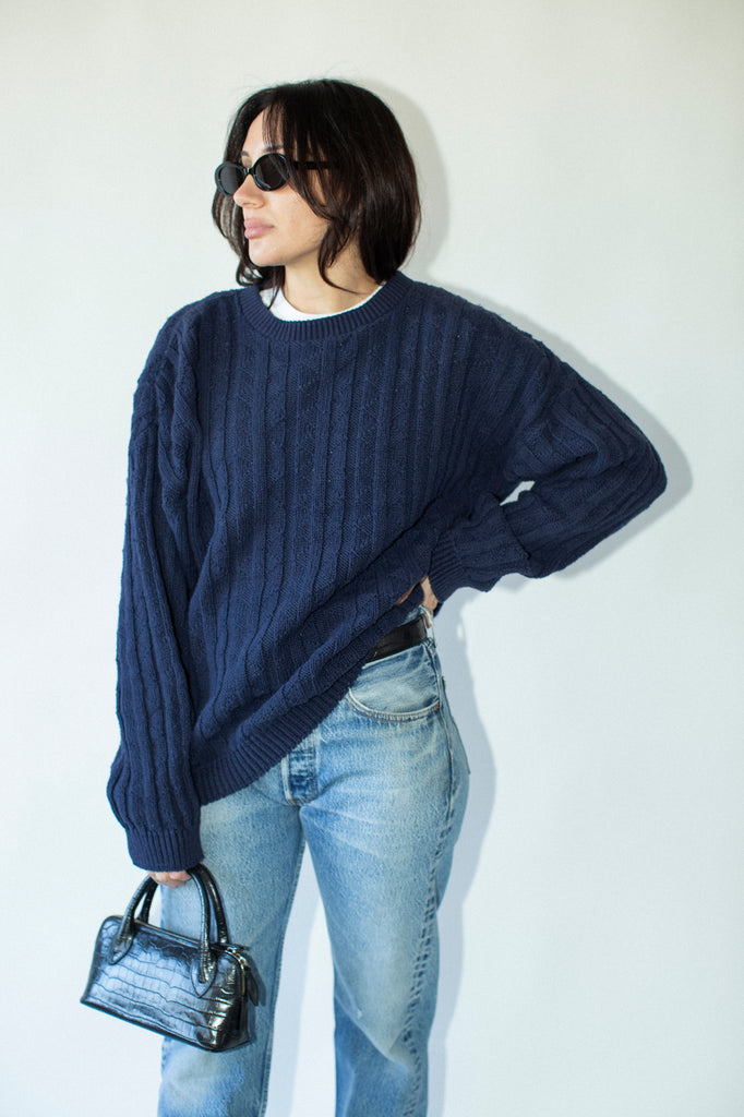 Person wearing a navy sweater and blue jeans, holding a black handbag against a white background