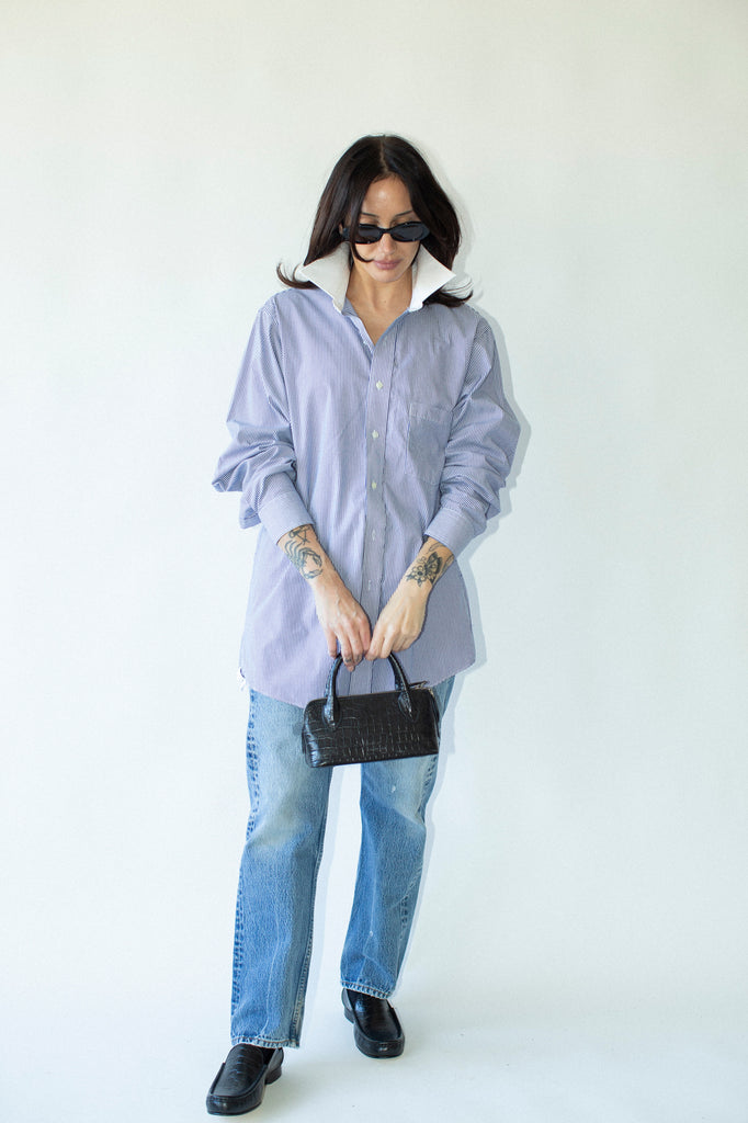 Person wearing a light blue oversized shirt, blue jeans, black shoes, and holding a black handbag on a white background.
