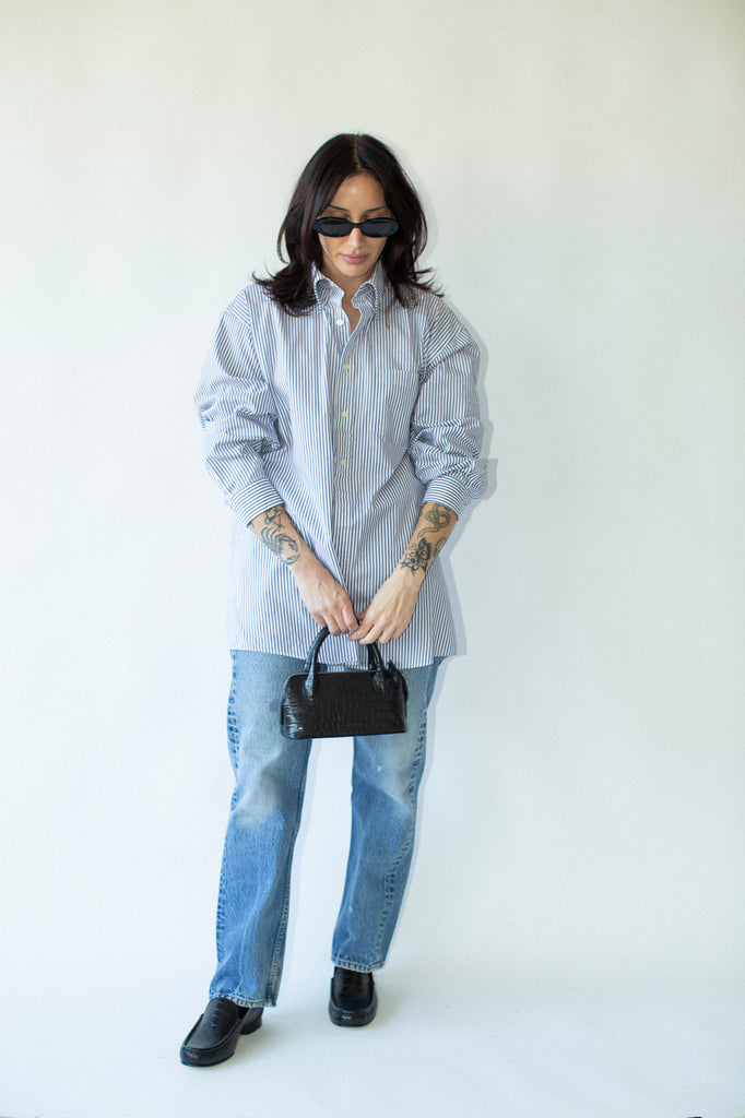 Person wearing a light blue shirt, jeans, and black shoes with a small black handbag on a white background