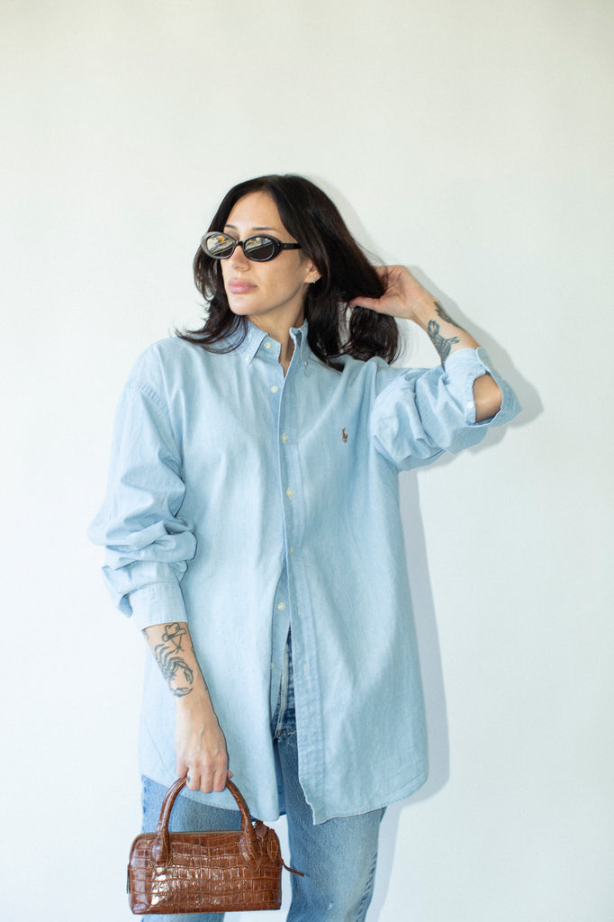 Woman wearing a light blue shirt and sunglasses, holding a brown handbag against a white background