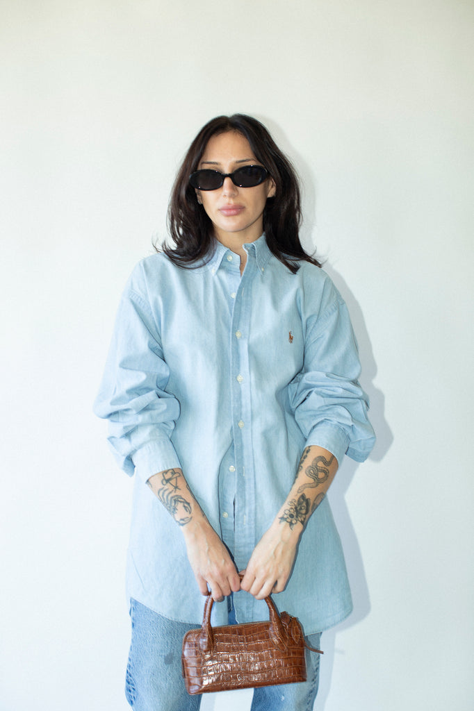 Woman wearing a light blue shirt and sunglasses, holding a brown handbag against a white background