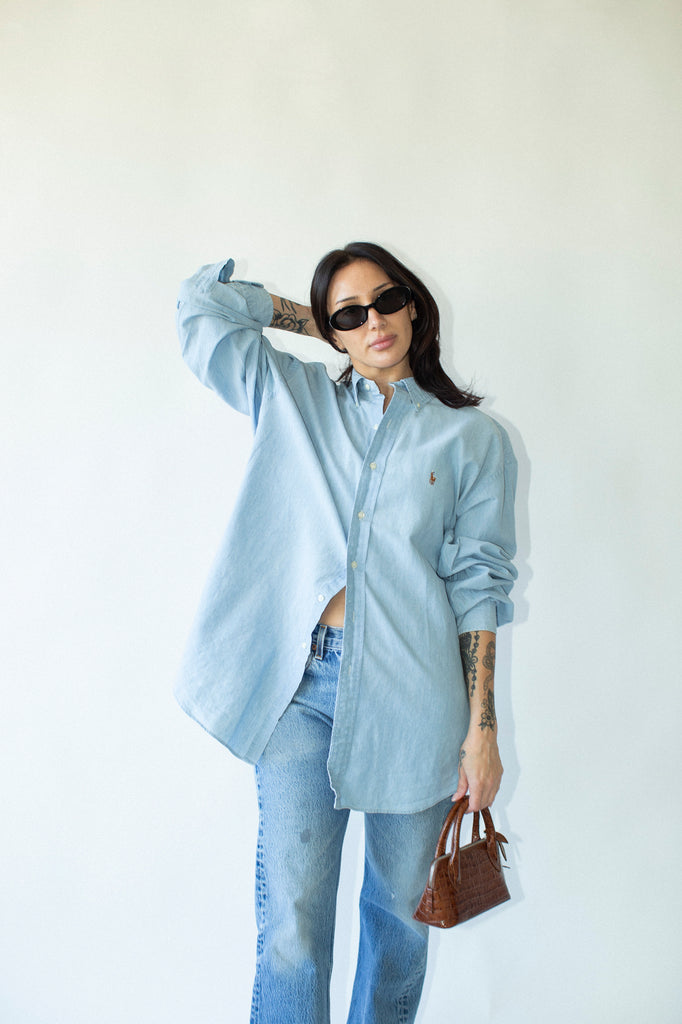 Person wearing a light blue oversized shirt and jeans, holding a brown handbag against a white background.