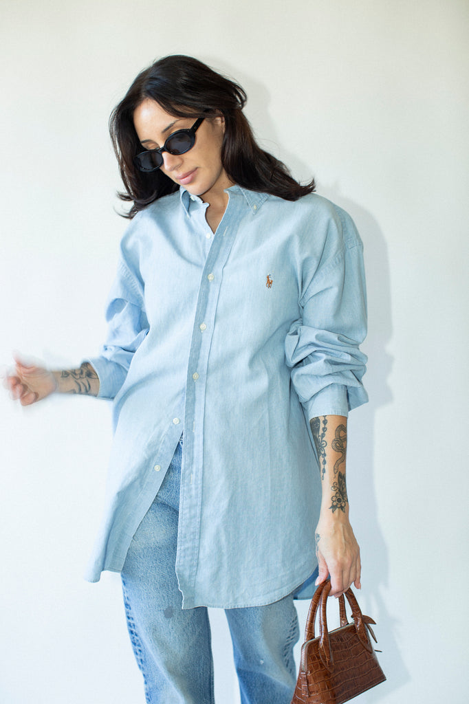 Woman wearing a light blue shirt with a logo, jeans, and sunglasses against a white background