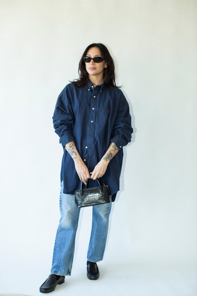 Person wearing a blue denim shirt and jeans, holding a black handbag against a white background