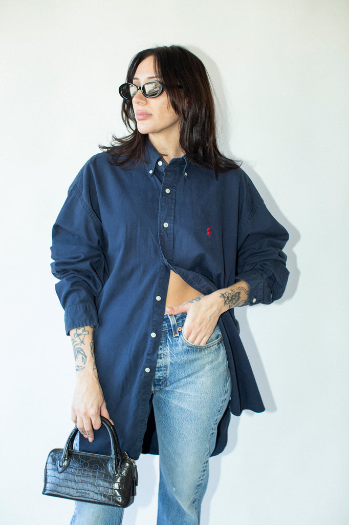 Woman wearing a blue shirt and jeans, holding a black handbag against a white background