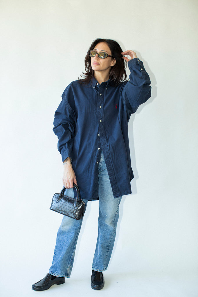Woman wearing a blue oversized shirt and jeans, holding a black handbag against a white background
