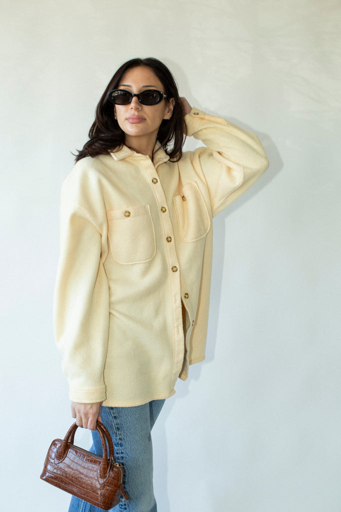 Woman wearing a light yellow jacket and sunglasses, holding a brown handbag against a white background