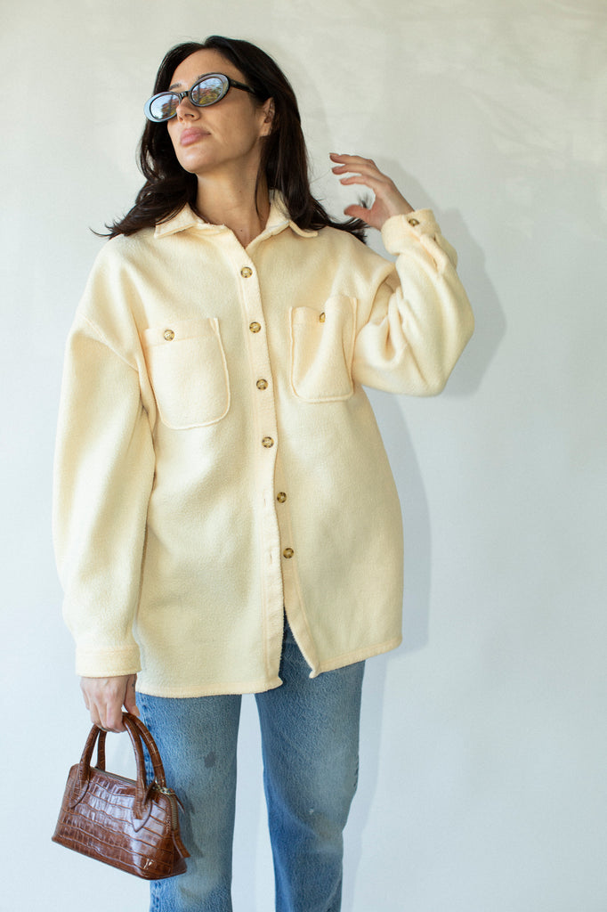 Woman wearing a light yellow jacket and blue jeans, holding a brown handbag against a white background