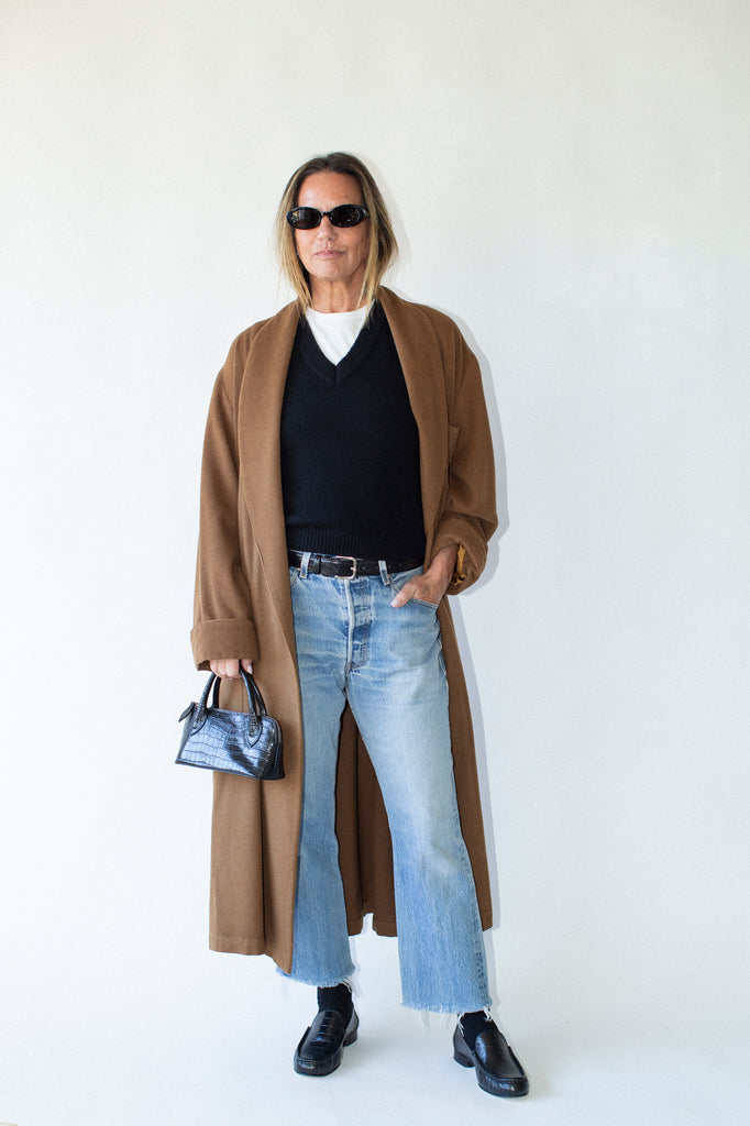 Person wearing a long brown coat, black shirt, light blue jeans, and black shoes on a white background.