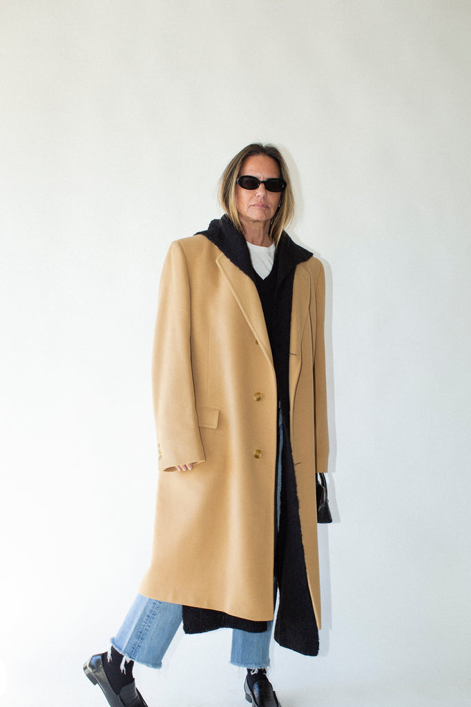 Person wearing a long beige coat over a black outfit with blue jeans against a white background