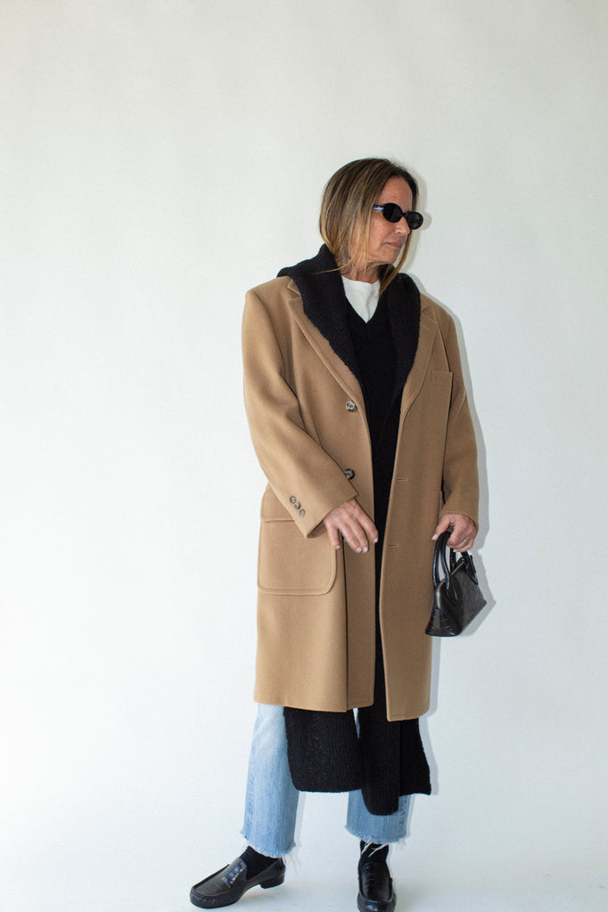 Person wearing a beige coat with a black scarf and sunglasses on a white background