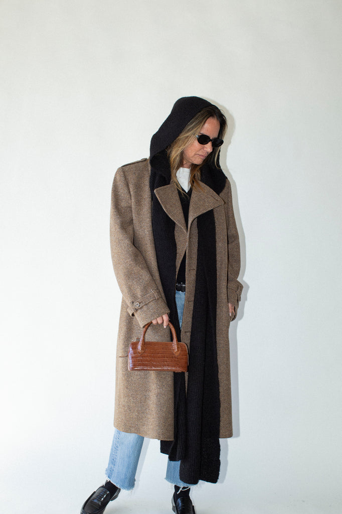 Person wearing a long brown coat with a hood, holding a brown handbag against a white background