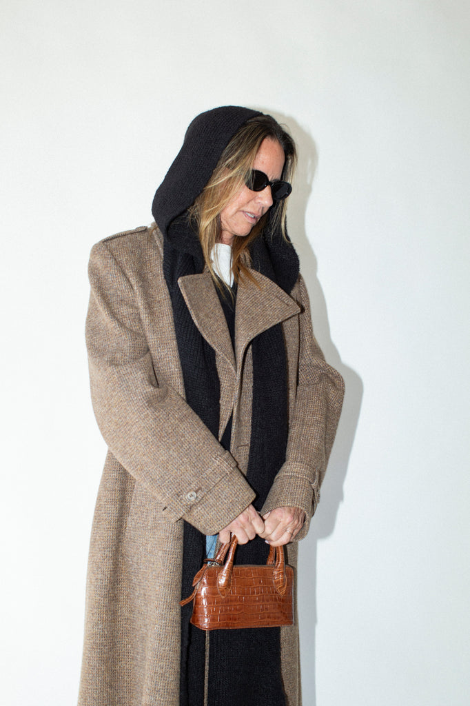 Person wearing a long beige coat and black hood, holding a brown handbag against a white background