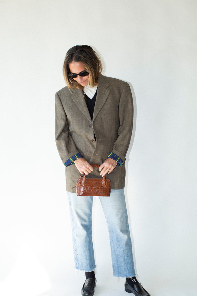 Person wearing a brown blazer and light blue jeans, holding a brown handbag against a white background.