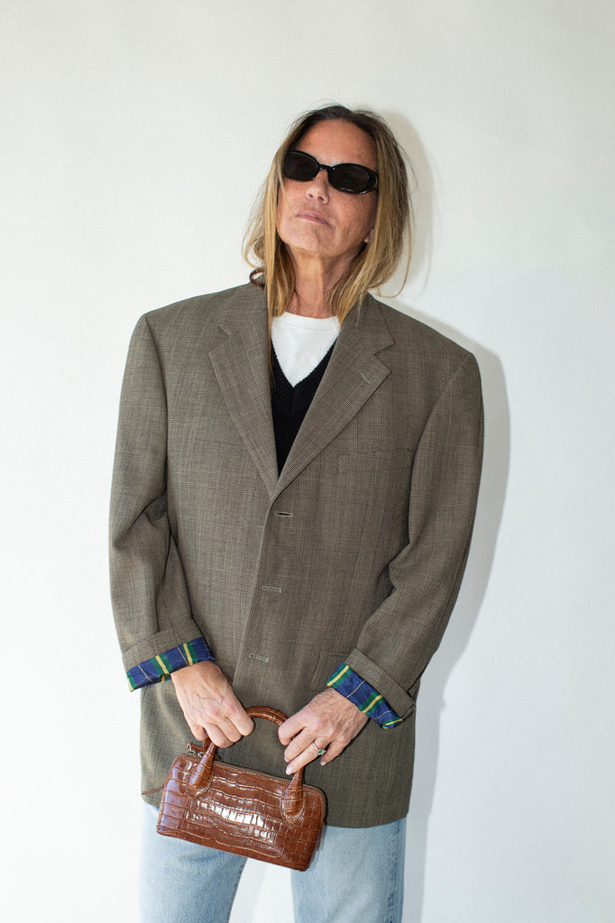 Person wearing a gray blazer and sunglasses, holding a brown handbag against a white background
