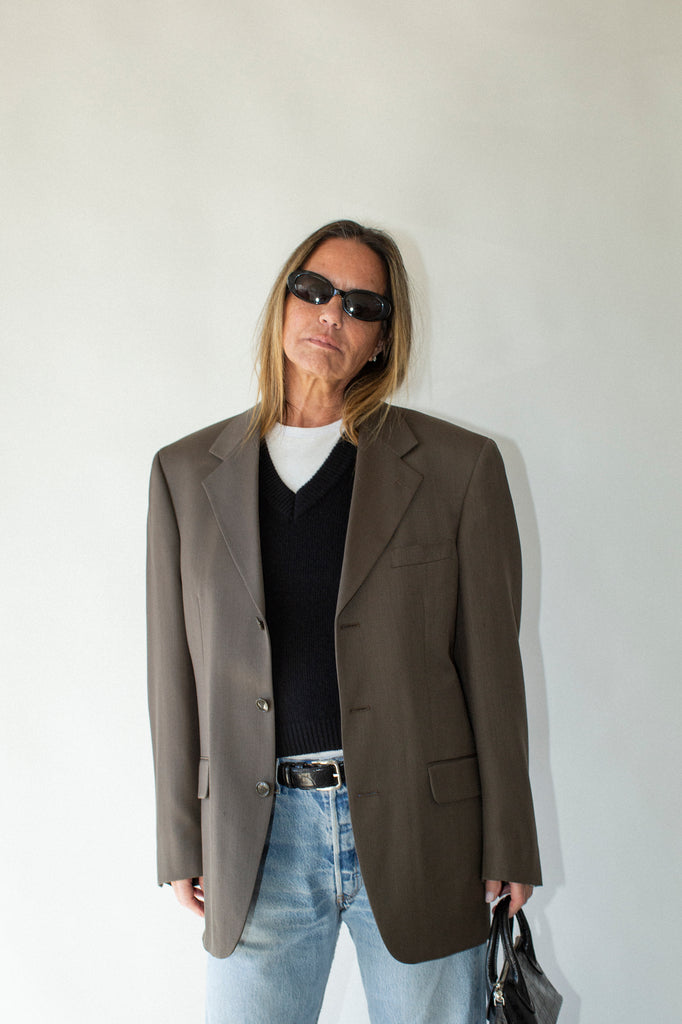 Person wearing a brown blazer, black shirt, and light blue jeans against a white wall.