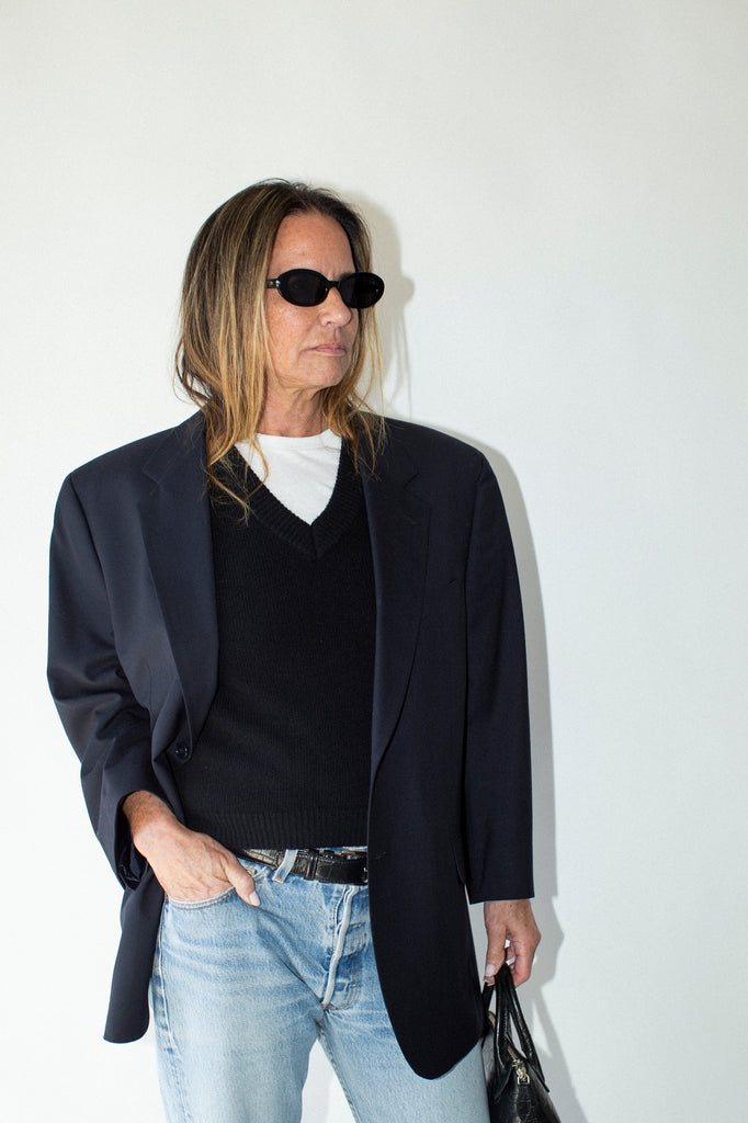 Person wearing a dark blazer, black shirt, light blue jeans, and sunglasses against a white background