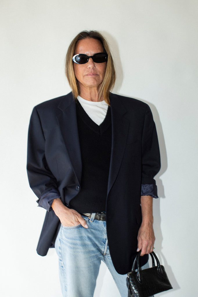 Person wearing a black blazer, black shirt, light blue jeans, and sunglasses, holding a black handbag against a white background.