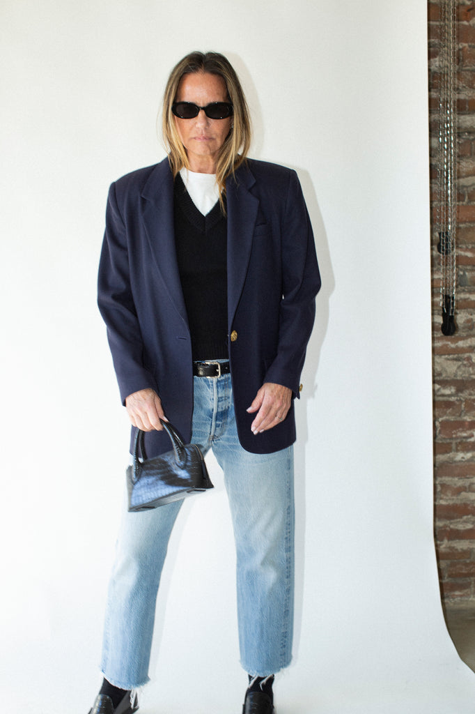 Person wearing a navy blazer, black shirt, light blue jeans, and sunglasses indoors.
