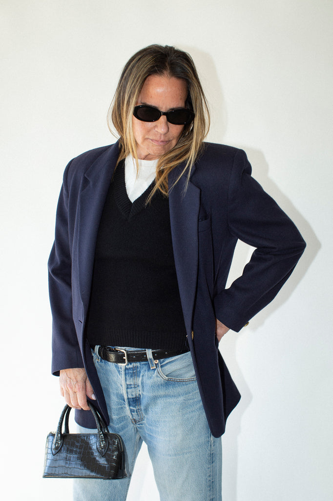 Person wearing a navy blazer, black top, light blue jeans, and sunglasses against a white background