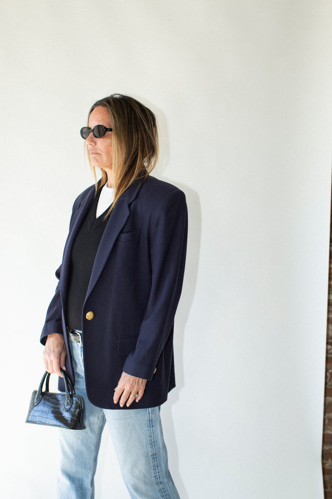 Woman wearing a navy blazer and sunglasses, standing against a white wall.