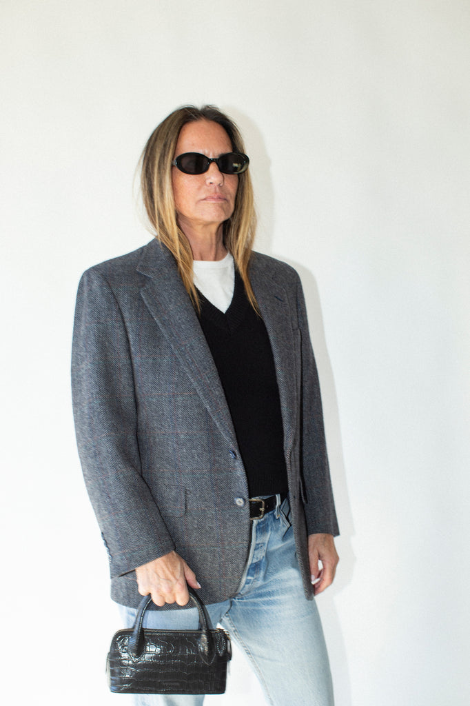 Person wearing a gray blazer, black top, light blue jeans, and sunglasses on a white background