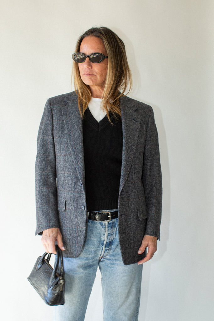 Person wearing a gray blazer, black shirt, light blue jeans, and sunglasses against a white background.