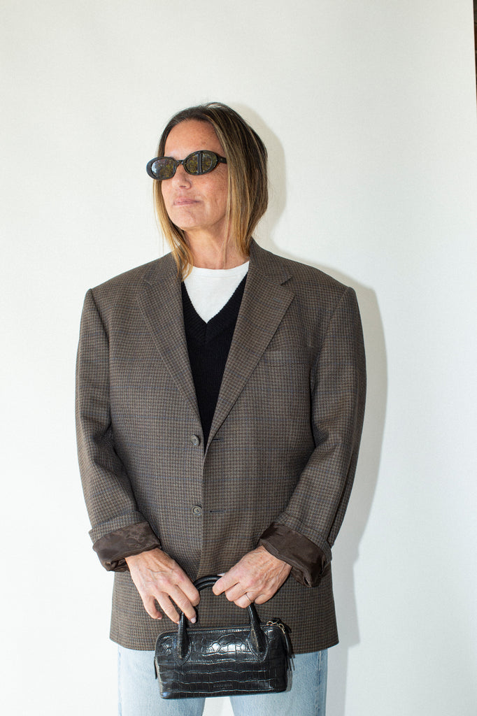Person wearing a brown blazer, black sunglasses, and holding a black handbag against a white background