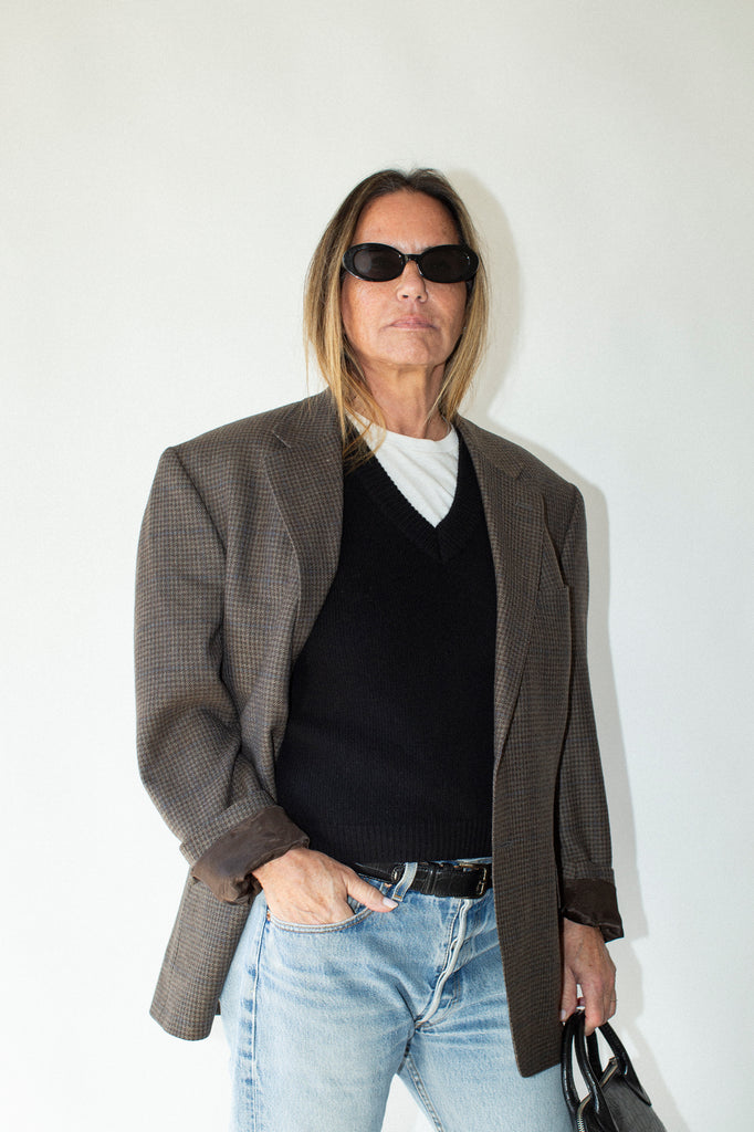Person wearing sunglasses, a brown blazer, black shirt, and light blue jeans against a white background