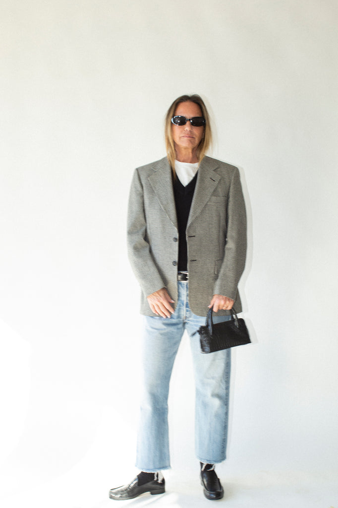 Person wearing a gray blazer, black shirt, light blue jeans, and sunglasses on a white background