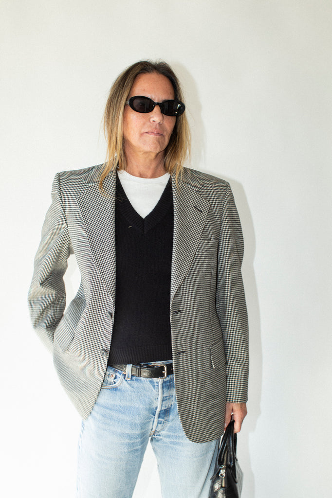 Person wearing a gray checkered blazer, black shirt, light blue jeans, and sunglasses against a white background.