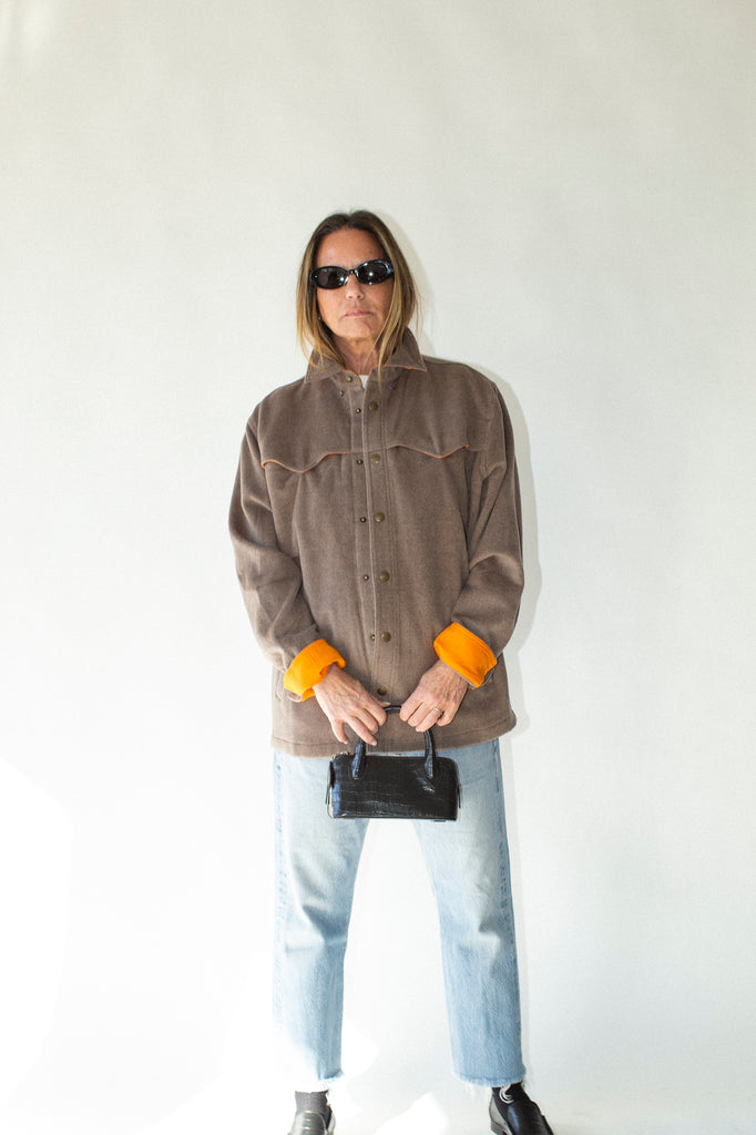 Person wearing a brown jacket with orange cuffs, light blue jeans, and black shoes against a white background.