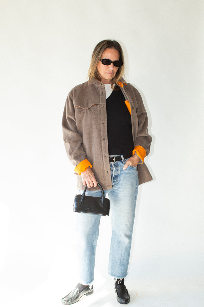 Person wearing a brown jacket with orange cuffs and collar, black shirt, light blue jeans, and sunglasses on a white background.