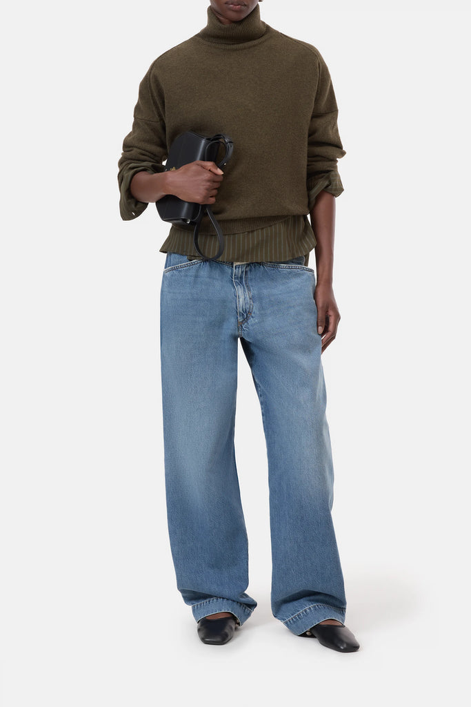 Person wearing a green sweater and blue jeans holding a black bag on a white background